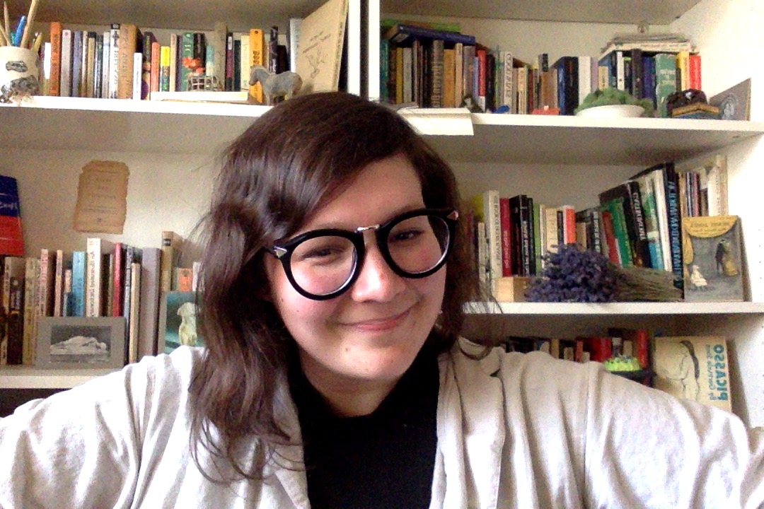 Alison Baitz self-portrait smiling at camera with black thick framed glasses in front of a bookshelf